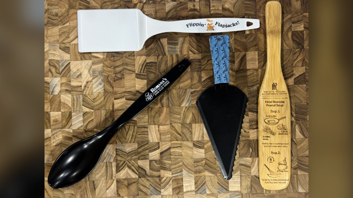 Four branded kitchen tools: an engraved wooden spatula, a black spoon with a one-color logo, and a turner and pie server with full-color prints