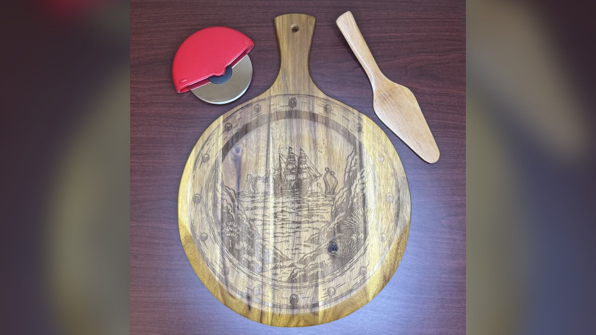 Round wooden pizza peel engraved with a sailing ship scene, with a red pizza cutter and a wooden server