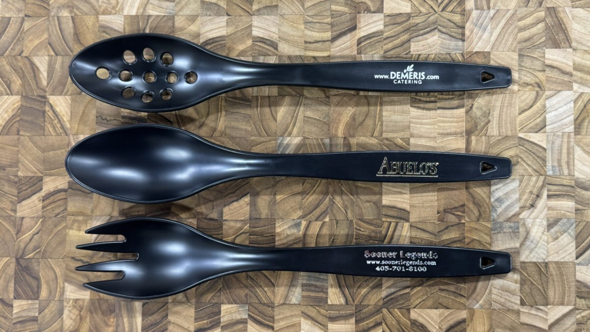 Three black serving utensils printed with company logos, laid on an end-grain wood board