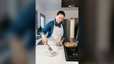 Smiling cook in a striped apron plating food with tongs at a kitchen counter