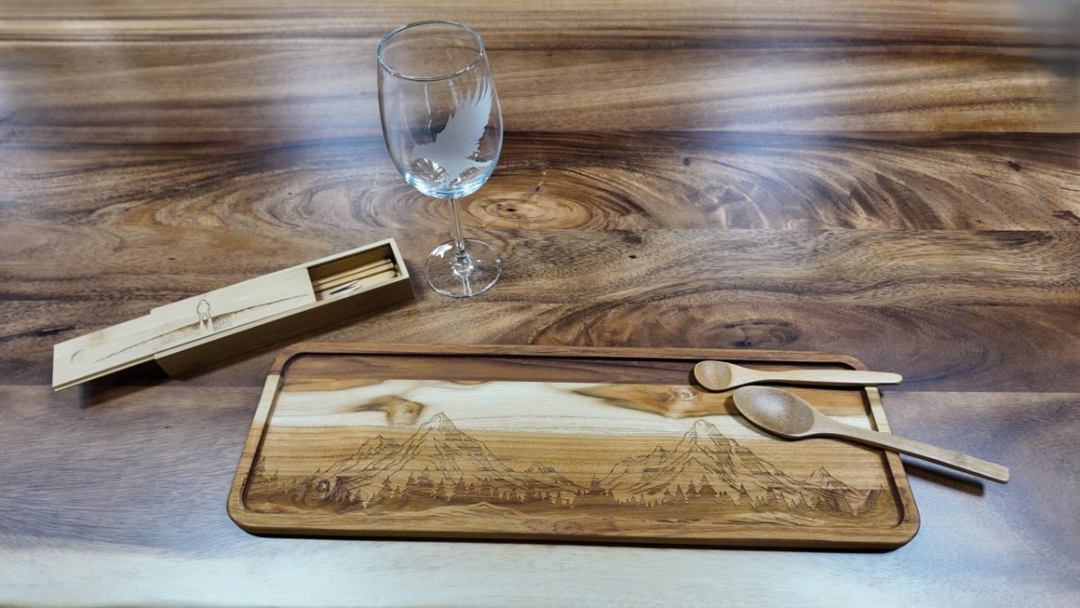 Engraved wooden serving tray with a mountain scene, two wooden spoons, a wooden box and an etched wine glass